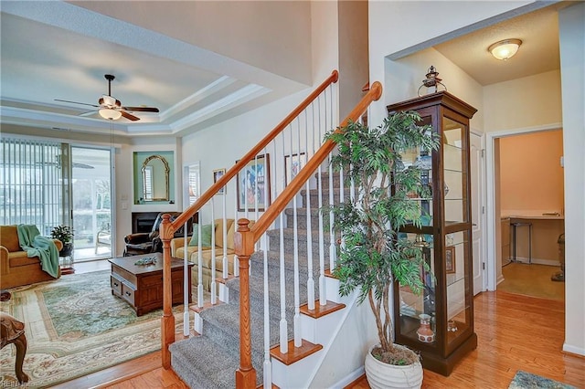 stairway featuring a fireplace, wood finished floors, a ceiling fan, a raised ceiling, and crown molding