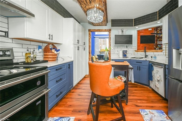 kitchen with a sink, wood finished floors, appliances with stainless steel finishes, blue cabinetry, and wall chimney exhaust hood