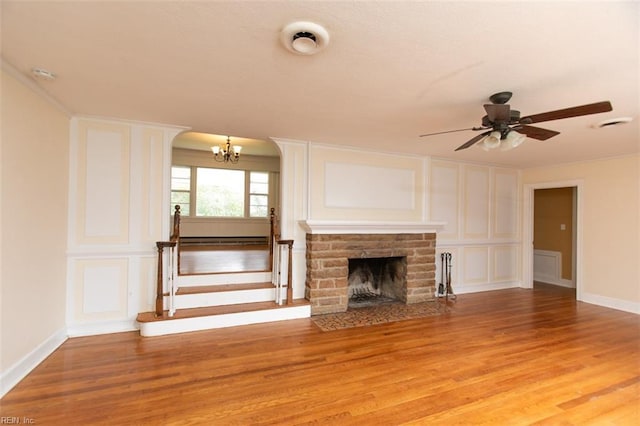 unfurnished living room featuring light wood-style flooring, a fireplace, stairs, a decorative wall, and ceiling fan with notable chandelier