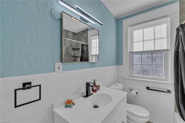 full bathroom with vanity, a wainscoted wall, tiled shower, toilet, and a textured wall