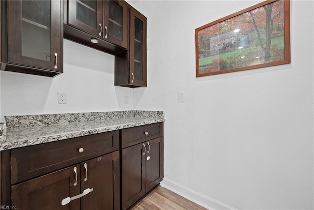 kitchen featuring light stone countertops, baseboards, dark brown cabinets, glass insert cabinets, and light wood-style floors