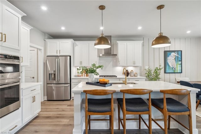 kitchen with wall chimney range hood, light wood-type flooring, light countertops, a kitchen breakfast bar, and appliances with stainless steel finishes