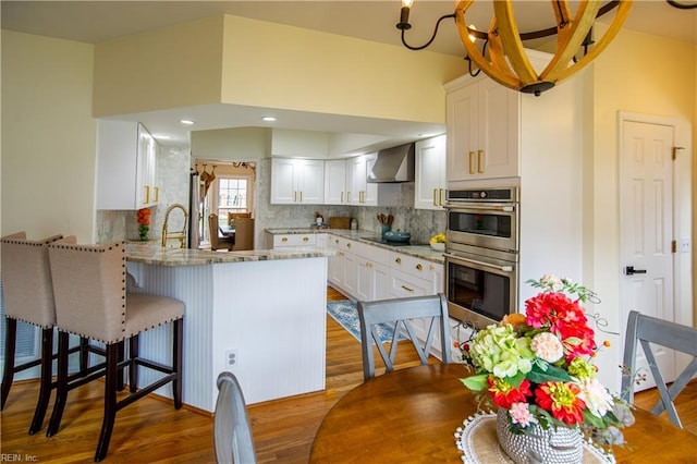 kitchen featuring backsplash, double oven, a peninsula, white cabinets, and wall chimney range hood