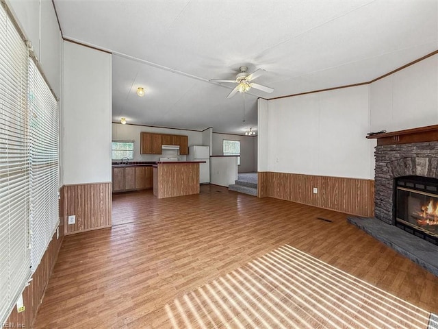 unfurnished living room with a ceiling fan, light wood-style floors, a stone fireplace, wood walls, and wainscoting