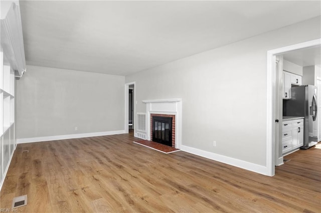 unfurnished living room with a brick fireplace, wood finished floors, visible vents, and baseboards