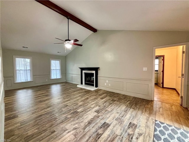 unfurnished living room featuring lofted ceiling with beams, wood finished floors, a glass covered fireplace, a decorative wall, and ceiling fan