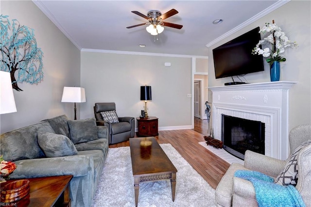 living room featuring a ceiling fan, wood finished floors, baseboards, crown molding, and a brick fireplace