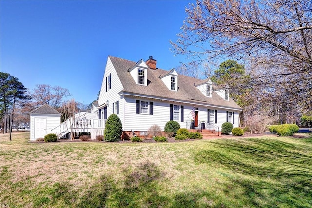 new england style home featuring an outbuilding, roof with shingles, a chimney, a front lawn, and a storage shed