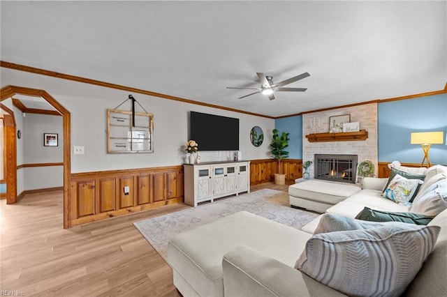 living area with a wainscoted wall, ceiling fan, light wood-style floors, crown molding, and a brick fireplace