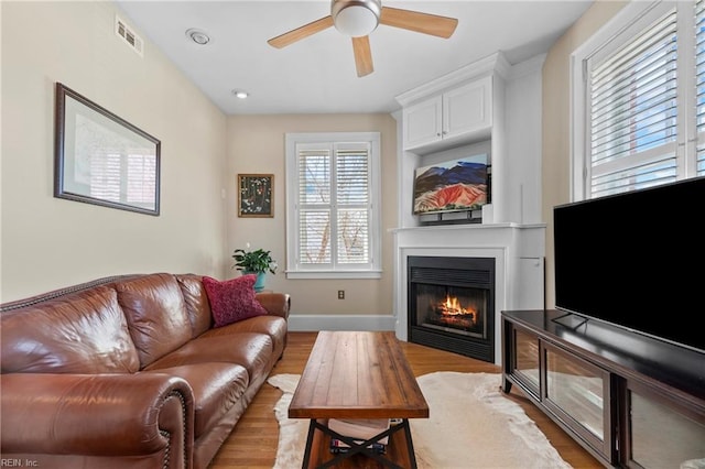 living area with visible vents, light wood-style flooring, a glass covered fireplace, baseboards, and ceiling fan