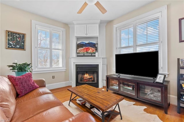 living area featuring light wood finished floors, ceiling fan, a lit fireplace, and baseboards