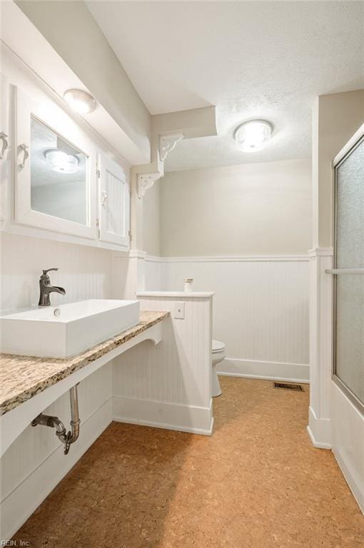 full bathroom with a shower with shower door, a wainscoted wall, visible vents, toilet, and a sink