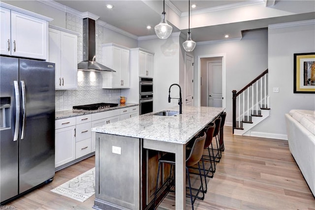 kitchen featuring light wood-type flooring, a kitchen island with sink, a sink, appliances with stainless steel finishes, and wall chimney range hood