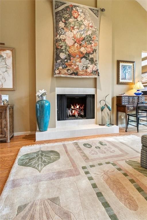 living area with baseboards, a warm lit fireplace, wood finished floors, and a towering ceiling