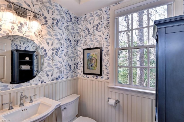 half bathroom with wallpapered walls, toilet, a sink, and wainscoting