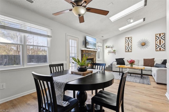 dining area featuring light wood-type flooring, vaulted ceiling with skylight, a fireplace, a textured ceiling, and a ceiling fan