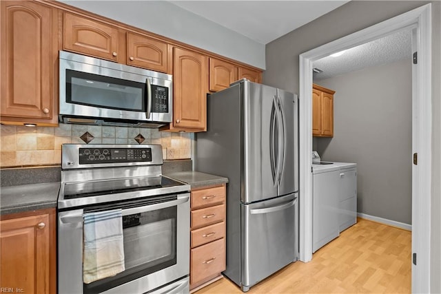 kitchen featuring light wood-style flooring, dark countertops, appliances with stainless steel finishes, decorative backsplash, and washing machine and clothes dryer