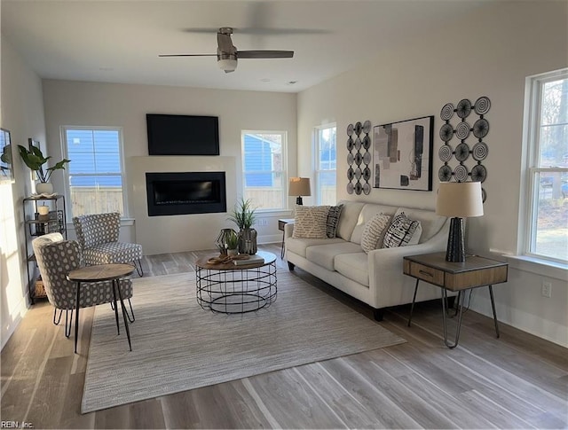 living area featuring a glass covered fireplace, wood finished floors, baseboards, and a ceiling fan