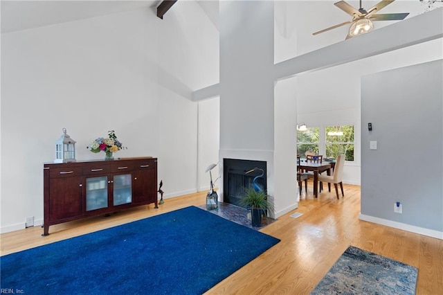 living room featuring wood finished floors, high vaulted ceiling, beam ceiling, a fireplace with flush hearth, and ceiling fan