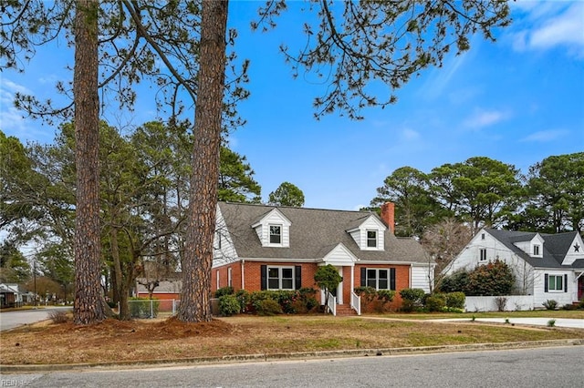 cape cod home featuring brick siding, a chimney, a front yard, and a shingled roof