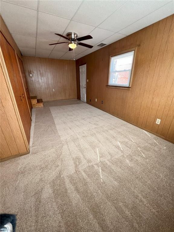 carpeted spare room with a drop ceiling, visible vents, wooden walls, and a ceiling fan