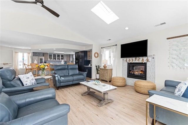 living area featuring a ceiling fan, visible vents, light wood finished floors, a premium fireplace, and lofted ceiling with skylight