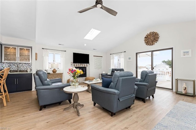 living area with vaulted ceiling with skylight, light wood-style flooring, a fireplace, and a ceiling fan