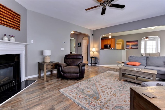 living area with arched walkways, a glass covered fireplace, ceiling fan, and wood finished floors