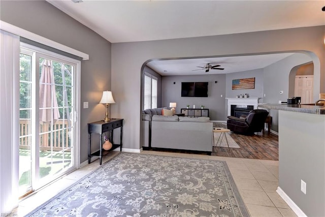 tiled living room with arched walkways, a fireplace, baseboards, and ceiling fan