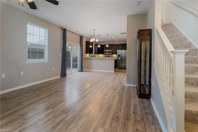 unfurnished living room featuring stairway, baseboards, a ceiling fan, and light wood finished floors