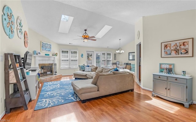 living area featuring a brick fireplace, baseboards, lofted ceiling with skylight, ceiling fan with notable chandelier, and light wood-style floors