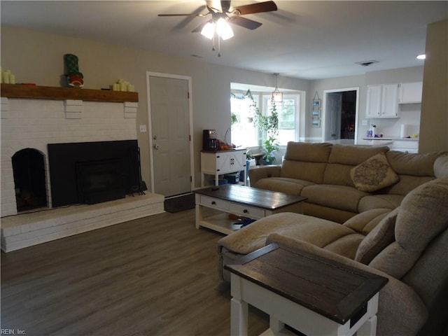 living room featuring a brick fireplace, dark wood-style floors, visible vents, and ceiling fan