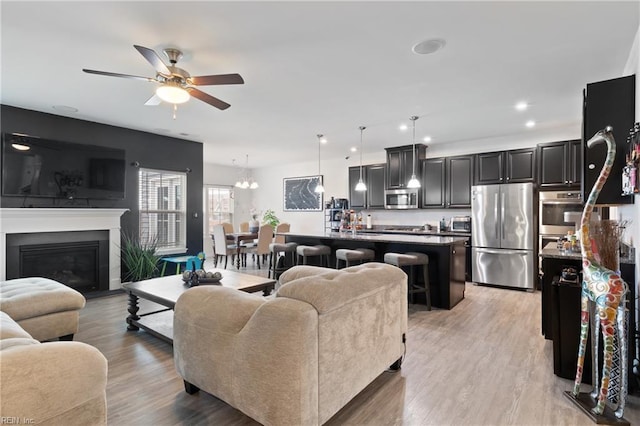 living room featuring a glass covered fireplace, recessed lighting, ceiling fan with notable chandelier, and light wood-style floors