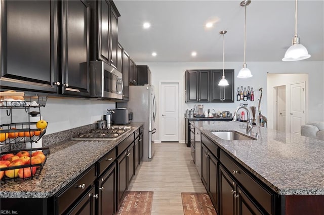 kitchen featuring dark stone counters, an island with sink, light wood-style floors, stainless steel appliances, and a sink