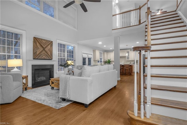 living area with a fireplace with flush hearth, light wood-style flooring, a towering ceiling, ceiling fan, and stairs