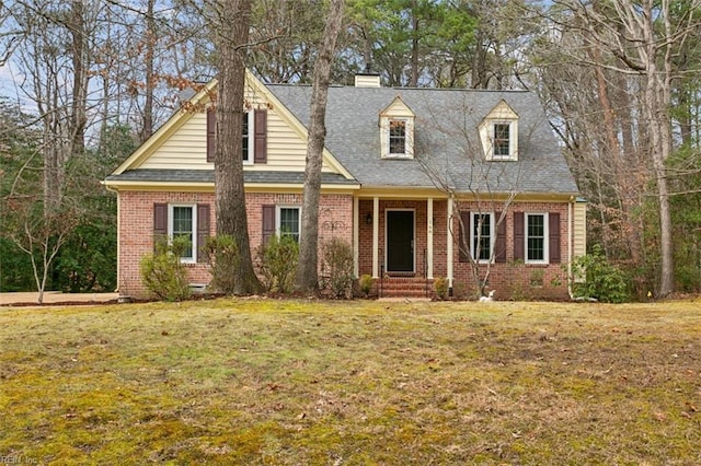 cape cod home with brick siding, a chimney, a front yard, and roof with shingles