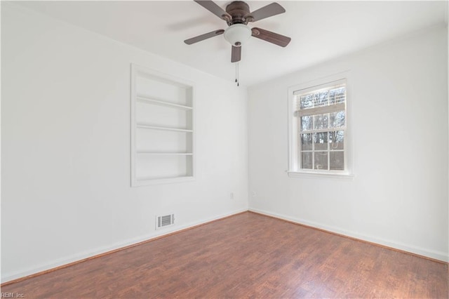 empty room with built in shelves, a ceiling fan, baseboards, and wood finished floors