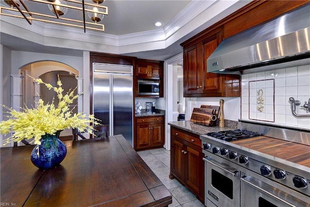 kitchen featuring wall chimney exhaust hood, high end appliances, a tray ceiling, and crown molding