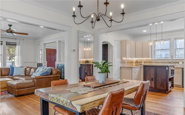 dining room with light wood-type flooring, decorative columns, ornamental molding, ceiling fan with notable chandelier, and arched walkways