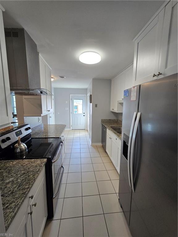 kitchen with light tile patterned floors, dark stone counters, white cabinets, appliances with stainless steel finishes, and wall chimney exhaust hood