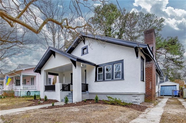 view of front of house featuring a shed, a porch, stucco siding, a chimney, and an outdoor structure