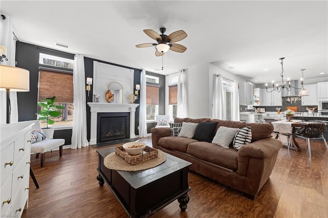 living room featuring a fireplace, ceiling fan with notable chandelier, dark wood-style floors, and visible vents