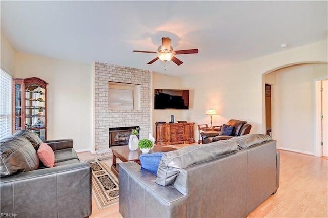 living room featuring a ceiling fan, arched walkways, a fireplace, light wood finished floors, and baseboards