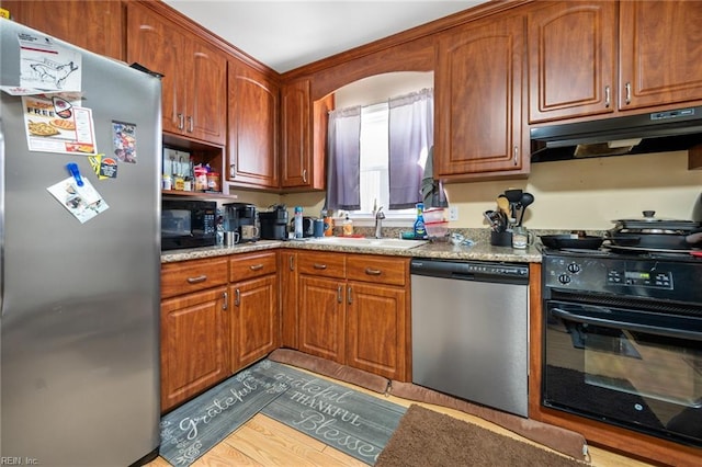 kitchen with under cabinet range hood, appliances with stainless steel finishes, wood finished floors, brown cabinetry, and a sink