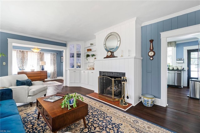 living room with built in features, wood finished floors, baseboards, a fireplace with raised hearth, and ornamental molding