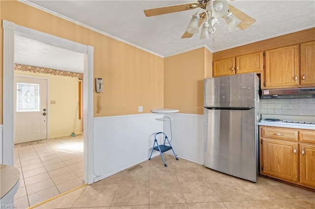 kitchen featuring a wainscoted wall, under cabinet range hood, freestanding refrigerator, light tile patterned flooring, and brown cabinetry