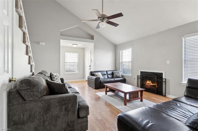 living area with light wood-style floors, a lit fireplace, baseboards, ceiling fan, and vaulted ceiling