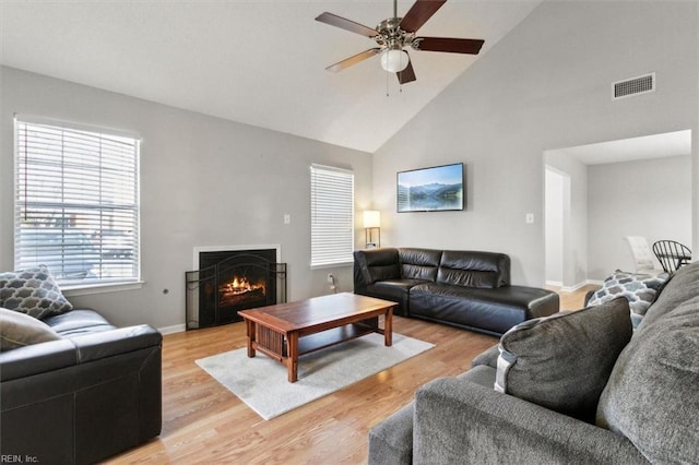 living room featuring visible vents, high vaulted ceiling, a ceiling fan, a warm lit fireplace, and light wood-style floors