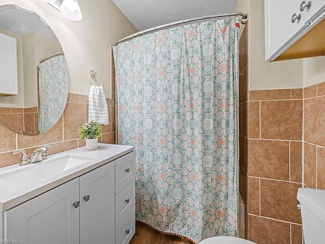 bathroom with toilet, vanity, wainscoting, a textured wall, and tile walls