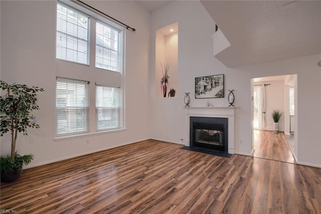 unfurnished living room featuring baseboards, a fireplace with flush hearth, wood finished floors, and a towering ceiling